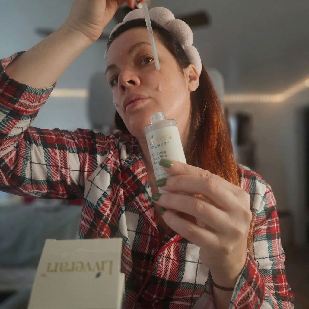 Woman applying cream to her face with a brush, wearing a plaid shirt indoors.