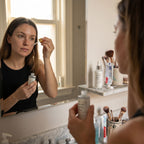 Woman applying skincare product in front of a bathroom mirror with various skincare items on the counter.